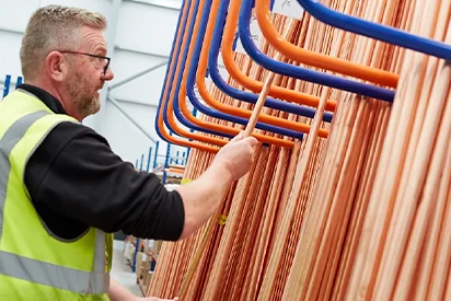 Male warehouse worker wearing a high-visibility vest selecting copper pipes from a vertical storage rack.