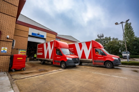 Two bright red delivery trucks with a bold white W wolseley are parked outside a trade supply warehouse. The warehouse has a blue sign above the entrance, and the surrounding area includes a concrete yard, trees, and a lamppost.